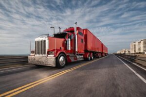 Red semi truck with a long enclosed trailer driving on a coastal highway under a cloudy sky, representing professional freight transportation and long-haul trucking services.