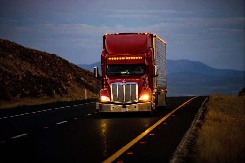 Red semi truck driving on a highway at night with headlights on and mountains in the background